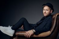 Der Sänger Johannes Oerding sitzt auf einem braunen Ledersessel. Er trägt einen schwarzen Anzug, einen schwarzen Hut und weiße Sneaker.