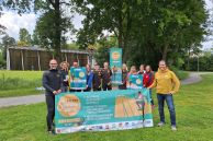 Das Foto zeigt eine große Gruppe aus Menschen, die sportlich gekleidet sind. Sie halten Banner, Plakate und Flyer zum &quot;Sälzer Sommer Sport&quot; in den Händen. Sie stehen auf einer grünen Wiese. Im Hintergrund sieht man Bäume und die Saline von Salzkotten.