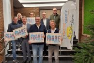 Acht Personen stehen in zwei Reihen hintereinander auf einer Treppe. Im Hintergrund sieht man eine rotgeklinkerte Wand und eine schwere Holztür. Bei den Personen handelt es sich um die Mitwirkenden des Adventskalenders der Stadt Salzkotten. Fünf Personen halten den Adventskalender in ihren Händen. Ein Mann rechts außen hält eine große, weiße Beachflag fest, die neben ihm steht, fest. Sie hat den Aufdruck Förderverein Sälzer Lagune.