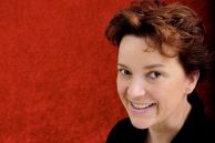 Autorin Kathrin Heinrichs hält einen Roman in der Hand und lächelt in die Kamera.