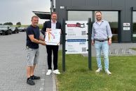 Im Hintergrund steht ein graues Firmengebäude mit grauer Fassade. Davor stehen Bürgermeister Ulrich Berger, Wirtschaftsförderer Peter Finke und Inhaber Frank Meyer. Bürgermeister Berger überreicht Herrn Meyer eine Urkunde der Stadt Salzkotten.