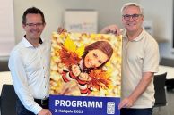 Zwei Männer stehen nebeneinander und halten ein Plakat mit dem Hinweis auf das neue Kursprogramm der VHS in den Händen