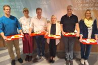 Sechs Personen, davon vier Männer und zwei Frauen, stehen in der Mensa der Gesamtschule Salzkotten. Jeder hält ein rotes Tablett in den Händen, auf dem ein Teller mit Essen angerichtet ist. Sie stehen nebeneinander vor einer Ausgabetheke in der Mensa.