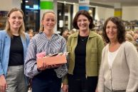 Vier Frauen stehen in den Räumlichkeiten der Stadtbibliothek. Eine von ihnen ist die Referentin. Sie hält eine lachsfarbene Kiste mit kleinen Geschenken in der Hand. Im Hintergrund sieht man die Besucherinnen der Veranstaltung.