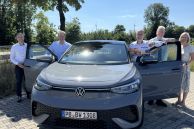 In der Mitte des Fotos steht ein grauer VW. Die vorderen Türen sind geöffnet. Hinter der linken Tür stehen zwei Männer und hinter der rechten Tür stehen zwei Männer und eine Frau.