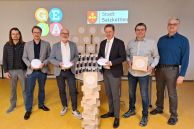 Fünf Männer stehen in einem Klassenraum. Vor ihnen ist ein Turm aus WLAN-Routern gestapelt. Hinter ihnen steht ein Tisch, auf dem weitere Router stehen. Im Hintergrund werden das Logo der Gesamtschule Salzkotten und das der Stadt Salzkotten mit einem Beamer an die Wand projiziert. Die Männer halten ebenfalls Router in den Händen. Drei von ihnen stehen links von dem Turm als Routern und drei rechts davon.