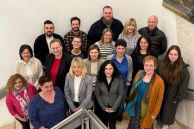 18 Personen stehen nebeneinander auf einer Treppe vor einer weißen Wand. Das Foto wurde von oben aufgenommen. Die Personen sind die Case Managerinnen und Manager des Kreises Paderborn.