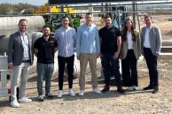 Sechs Männer und eine Frau stehen vor einem Bauzaun. Dahinter sieht man eine Baustelle. Eine Erdhaufen türmen sich und links ist ein Bagger zu sehen. Über den Personen ist ein großes Schild aufgestellt, auf dem &quot;Neubau eines Logistikzentrums mit Bürofläche, ARNOWA und Goldbeck&quot; steht. Eine Animation des entstehenden Gebäudes ist ebenfalls zu sehen.