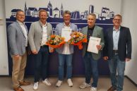 Fünf Männer stehen im Foyer des Rathauses Salzkotten. Hinter ihnen steht ein großer Aufsteller, der die Silhouette von Salzkotten zeigt. Zwei von ihnen halten Urkunden und Blumensträuße in den Händen.