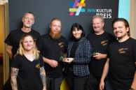 Sechs Personen stehen vor einem hohen Aufsteller, auf dem &quot;WW Kulturpreis 2025&quot; steht. Fünf Personen gehören zum Rock Musik Verein. Sie tragen schwarze Shirts mit dem Logo des Vereins. In der Mitte steht ein Mann mit kurzen roten Haaren und einem schulterlangen roten Bart, der den Kulturpreis 2025 von einer Frau mit schwarzen, schulterlangen Haaren entgegennimmt. 