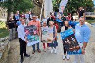 an die 30 Personen stehen als Gruppe nebeneinander vor dem Gradierwerk in Salzkotten. Sie halten Plakate zu den einzelnen Programmpunkten des Hederauenfests 2025 hoch. Vorne rechts in ein Oldtimer-Trecker zu sehen, auf dem einige Personen sitzen und stehen. Hinten in der Mitte des Bildes steht ein Oldtimer Audi.