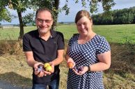 Frau Hagelganz und Herr Seyfzadeh stehen nebeneinander und halten frisch gepflückte Pflaumen und Äpfel in ihren Händen. Im Hintergrund sind Pflaumen- und Apfelbäume zu sehen, die am Straßenrand stehen