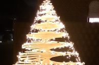 Ein Tannenbaum aus Holz. Die einzelnen Holzbretter sind an einem Stamm in der Mitte kreisförmig angelegt, sodass sich daraus ein gefächerter Baum ergibt. Auf der Spitze ist ein leuchtender Stern und der gesamte Baum ist mit einer Lichterkette umhüllt.