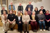 14 Personen sitzen auf einem Sofa oder stehen dahinter. Sie stehen vor einer beigen Wand, auf der "Kulturscheine 1a" steht.