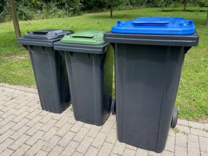 Drei Mülltonnen stehen nebeneinander an der Straße. Zwei Tonnen sind kleiner. Eine hat einen grauen Deckel und die andere einen grünen. Die Tonne ganz rechts ist größer und hat einen blauen Deckel.