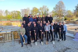 Mitglieder der Löscheinheit Upsprunge, die Beteiligten der Stadt Salzkotten, der Bauunternehmer und der Ortsvorsteher von Upsprunge stehen auf der Baustelle des Neubaus vom Feuerwehrgerätehaus in Upsprunge. Die Personen stehen in drei Reihen hintereinander. Die hintere Reihe steht erhöht auf einer aus Beton gegossenen Außenmauer. Die zwei vorderen Reihen stehen davor. Die Personen aus der vordersten Reihe halten alle einen Spaten in der Hand und deuten an, diesen in den Boden zu stemmen.