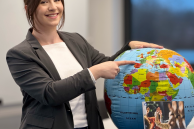 Eine Frau mit braunen zusammengebundenen Haaren steht neben einem Globus und zeigt auf Portugal. Im Vordergrund steht das neue VHS-Programm