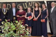 Bürgermeister Ulrich Berger, Volksbankvertreter Martin Herbst, Sofia Sabezki, Anna Kleine, Pia Isekenmeier, Nadja Aleinik, Schulleiter Berthold Fischer stehen auf der Bühne vor eine Pflanze.