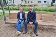 Zwei Männer sitzen nebeneinander auf einer großen Hollywoodschaukel, auf der insgesamt sechs Erwachsene Platz finden. Die Schaukel steht in einem Kiesbett. Dahinter läuft ein Gehweg entlang. Neben dem Gehweg ist ein Grünstreifen mit höheren Büschen und Efeu angelegt. Im Hintergrund sind drei Einfamilienhäuser zu sehen.