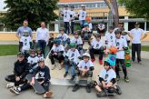 Eine Gruppe aus Kindern und Erwachsenen steht auf einer Skateanlage. Die Kinder tragen Helme und T-Shirts, die ein Skateboard drauf gedruckt haben.