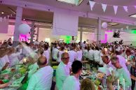 In einer großen Halle mit weißer Decke und weißen Säulen sitzen hunderte Menschen an nebeneinander aufgereihten Biertischgarnituren. Alle Personen trage ausschließlich weiße Kleidung. Die Tische sind mit weißer Deko und verschiedenem Essen und Getränken gedeckt. Durch Strahler wird die Halle in ein pinkes und grünes Licht getaucht. An der Decke hängen weiße Wimpelketten und an den Tischen sind große, schwebende, weiße Ballons befestigt.