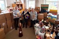 Drei Frauen stehen in einem Raum, der mit Kartons und Weihnachtsdekorationen voll gestellt ist. Die drei Frauen halten Weihnachtsdekoration in die Kamera