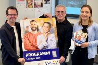 Drei Mitarbeiter der VHS präsentieren das neue Programmheft und halten ein Plakat hoch
