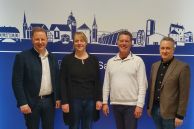 Ulrich Berger (Bürgermeister Stadt Salzkotten), Miriam Mattiza (Fachdienst 1.3), Rainer Vidal (Leiter DigitalBüro OWL) und Michael Rölleke (Leiter Fachbereich Zentrale Dienste) stehen vor der Silhouette der Stadt Salzkotten im Rathaus.
