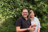 Sofia Hagelganz und Sam Seyfzadeh stehen an der Straße vor einem Pflaumenbaum und halten Obst in der Hand.