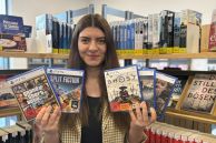 Eine Frau steht vor einem Bücherregal in der Stadtbibliothek Salzkotten. Sie hält fünf verschiedene Playstation-5-Spiele in den Händen