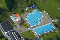Das Salzkottener Freibad aus der Vogelperspektive. Rechts sind zwei große Schwimmbecken zu sehen, links ein kleineres mit einem kleinen Turm mit einer Rutsche daran. Links ist ein längliches Gebäude. Das Gelände ist von grünen Wiesen umgeben.