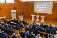 Auf dem Foto sieht man einen großen Saal. Vorne steht ein Mann, der einen Vortrag hält. Rechts neben ihm wird eine Präsentation an die Wand projiziert. Die angezeigte Folie zeigt die Aussage "Hass im Netz ist ein Problem. Strafrecht ist ein Teil der Lösung."